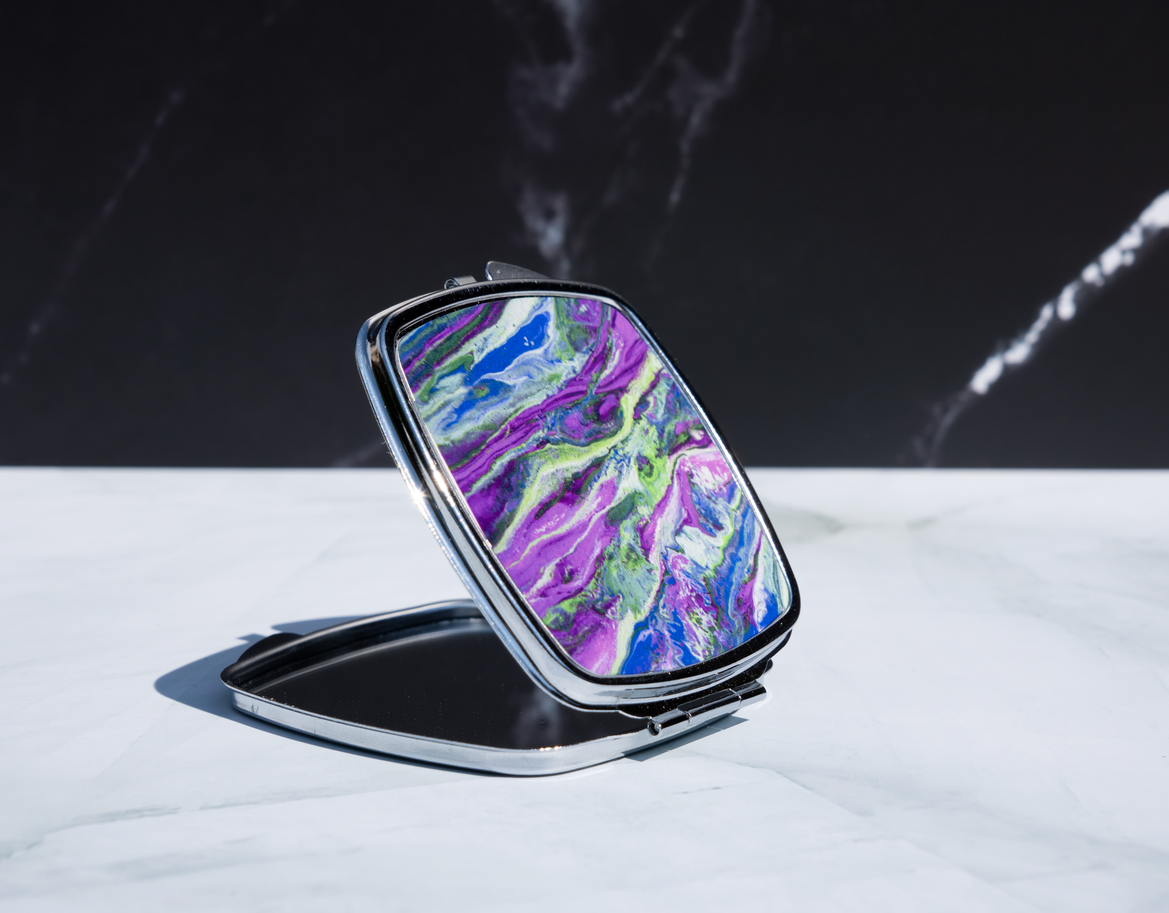 Square personalised compact mirror with abstract paint pour artwork on a metal case, displayed open on a marble background.